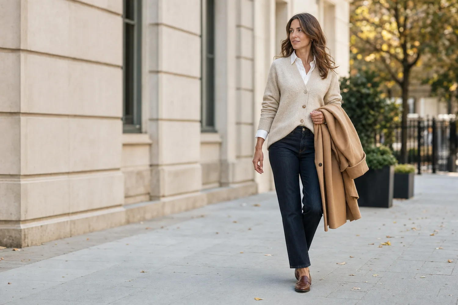 Woman wearing an oatmeal cardigan over a white collared shirt with dark jeans, brown loafers, and a camel coat